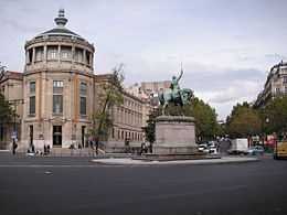 Photographie en couleur d'une place ornée d'une statue de cavalier, à gauche un immeuble en rotonde, à droite une avenue