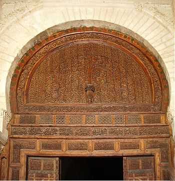 Photographie de la partie supérieure de la grande porte de la salle de prière. Le décor sculpté, notamment celui du tympan, comprend des éléments végétaux, géométriques et épigraphiques.