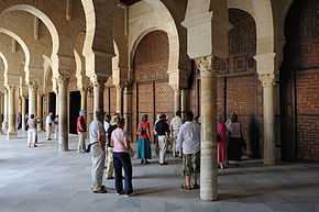 Photographie d’un groupe de touristes dans le portique à double travée, précédant la salle de prière. Celle-ci est entièrement fermée, côté portique, par des portes en bois sculpté.