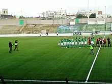 Dans un stade de football, des photographes vont sur le terrain pour photographier une équipe au maillot vert.