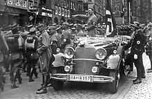 Photo noir et blanc prise en novembre 1935. Dans une rue de Nuremberg, Adolf Hitler debout à l’arrière d’une voiture au premier plan, honore, d’un salut nazi, un défilé de troupes de miliciens de la SA se déroulant sur sa gauche. On distingue aussi, à l’arrière de la voiture, juste derrière Hitler, le drapeau à croix gammée de cérémonie, Blutfahne, et, en uniforme SS, debout à droite, devant la portière de la voiture, Jakob Grimminger, porteur officiel du Blutfahne.