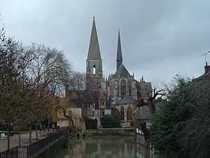 Paysage d'hiver, arbres partiellement dénudés, avec une église gothique, chœur avec arcs-boutants toit en ardoise, surmontée d'une flèche pointue recouverte aussi d'ardoises, et un clocher conique en pierre ; au premier plan un bassin d'eau verte