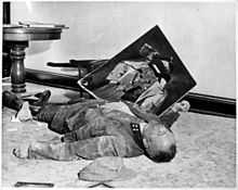 Photo noir et blanc prise le 19 avril 1945, à l’intérieur d’un bureau de l’hôtel de ville de Leipzig. Sur le sol, un homme mort, en uniforme, est allongé de tout son long, sa tête vers la droite au premier plan. Un mur blanc forme l’arrière-plan de la photo. Au second plan, entre une chaise renversée le long du mur et le flanc droit du cadavre, tient en équilibre un portrait d’Adolf Hitler dont le visage a été lacéré.